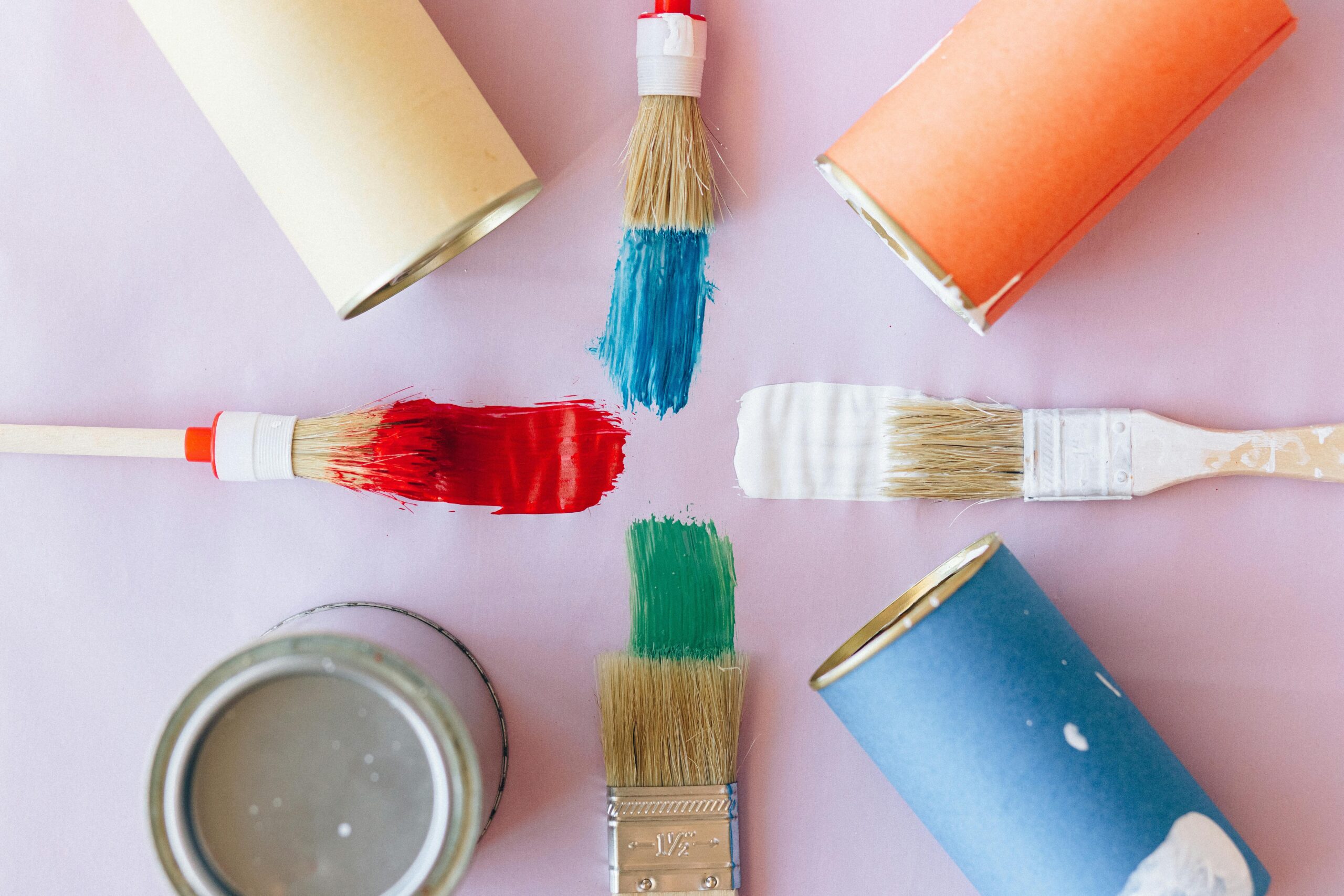 Four paintbrushes with red, blue, green, and white paint are arranged in a cross pattern on a light background, surrounded by open paint cans in matching colors.